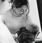 A close-up of a detailed graphite drawing showing a smiling mother in a ribbed turtleneck sweater gently looking down at the baby she is holding in her arms. The baby, wearing a dark knitted onesie, is lying on its back and looking up at its mother. The drawing conveys the love and bond between them.