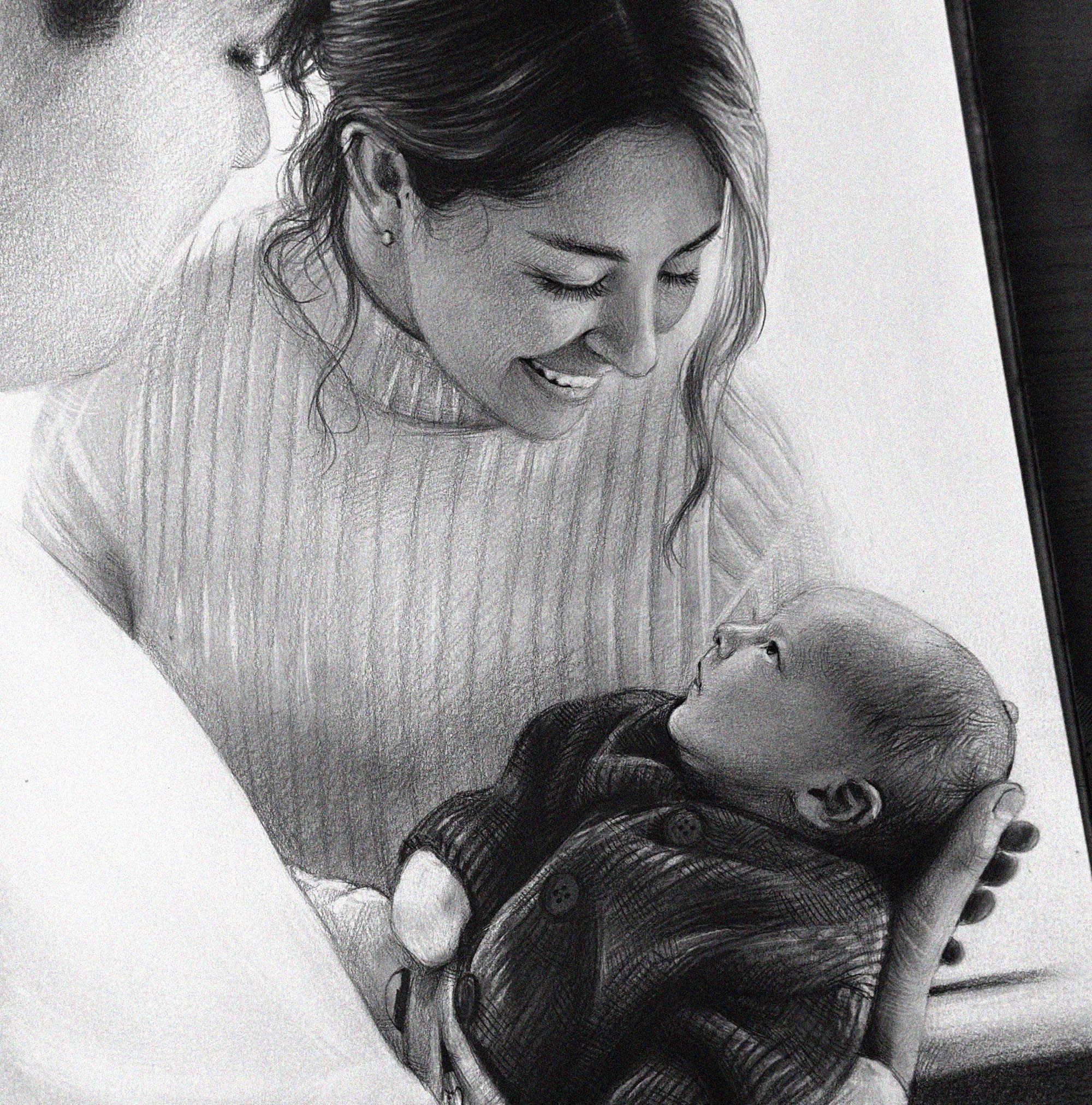 A close-up of a detailed graphite drawing showing a smiling mother in a ribbed turtleneck sweater gently looking down at the baby she is holding in her arms. The baby, wearing a dark knitted onesie, is lying on its back and looking up at its mother. The drawing conveys the love and bond between them.