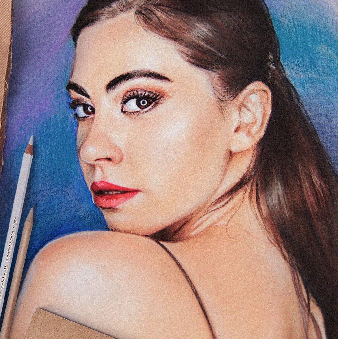 A close-up colored pencil portrait drawing of a woman with dark brown hair, red lips, and a blue background, with pencil and paper visible at the edges.
