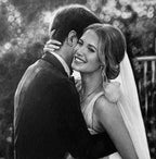 Detailed black and white graphite pencil drawing of a bride and groom embracing. The smiling bride is wearing a veil and her wedding ring is visible as she hugs the groom.
