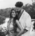 Detailed black and white graphite pencil drawing of a happy couple embracing outdoors. The smiling woman wears a jeweled headpiece and holds a bouquet, while the bearded man in a light jacket looks down at her affectionately.